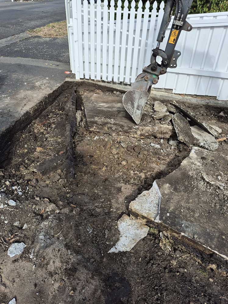 Excavator breaking up old concrete beside a white picket fence