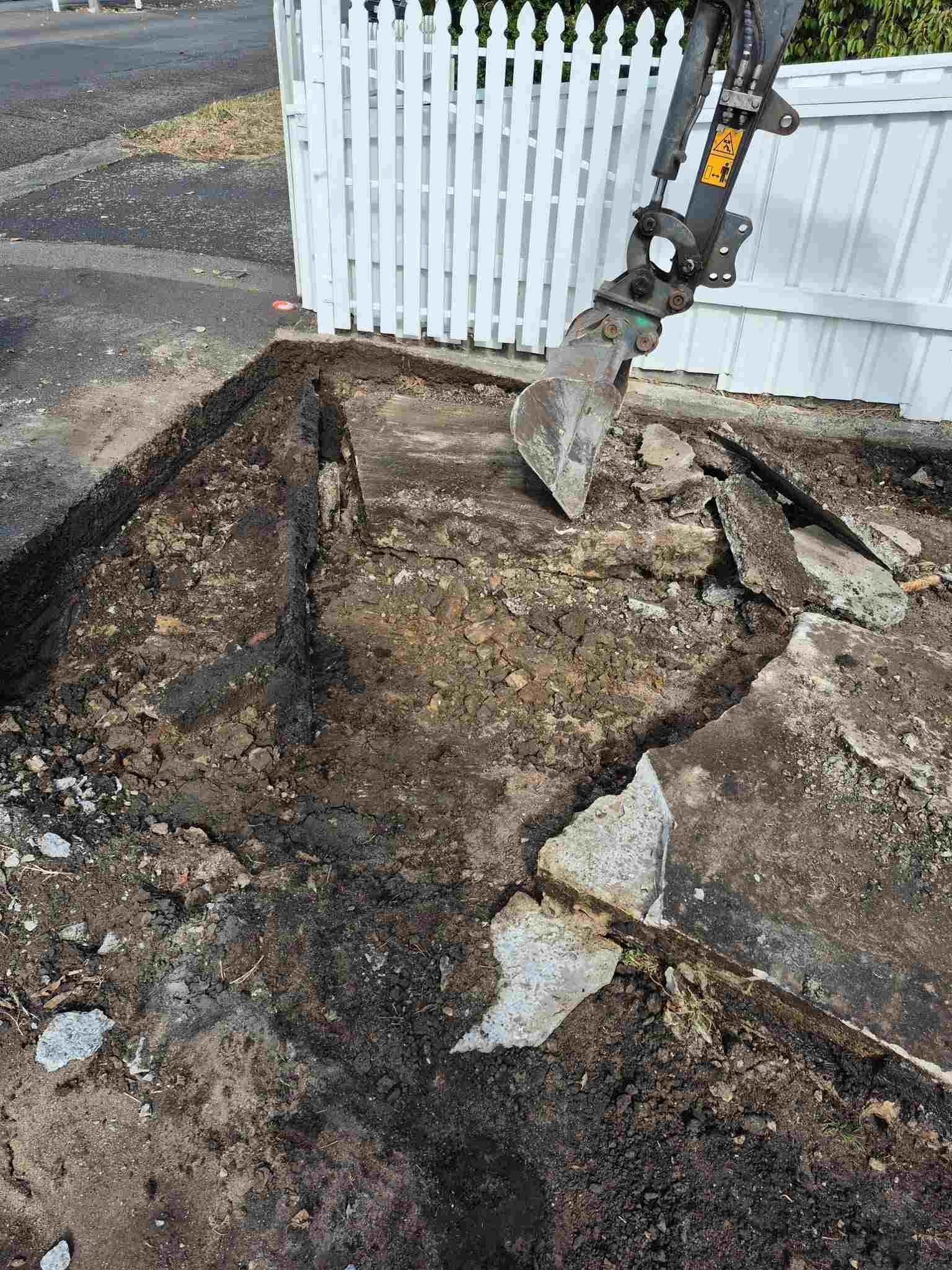 Excavator breaking up old concrete beside a white picket fence