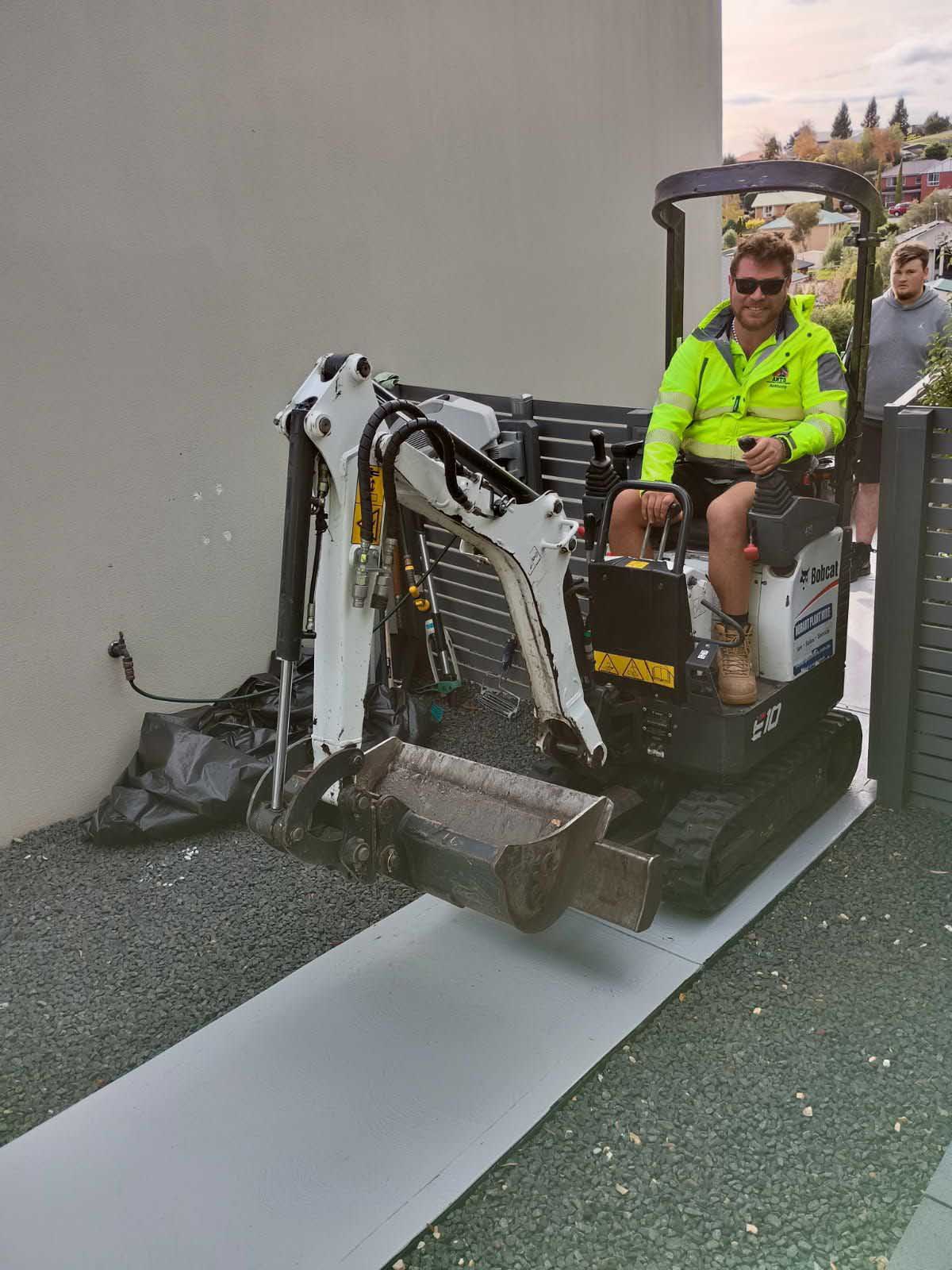 Man in hi-vis driving a mini excavator on a board beside a house
