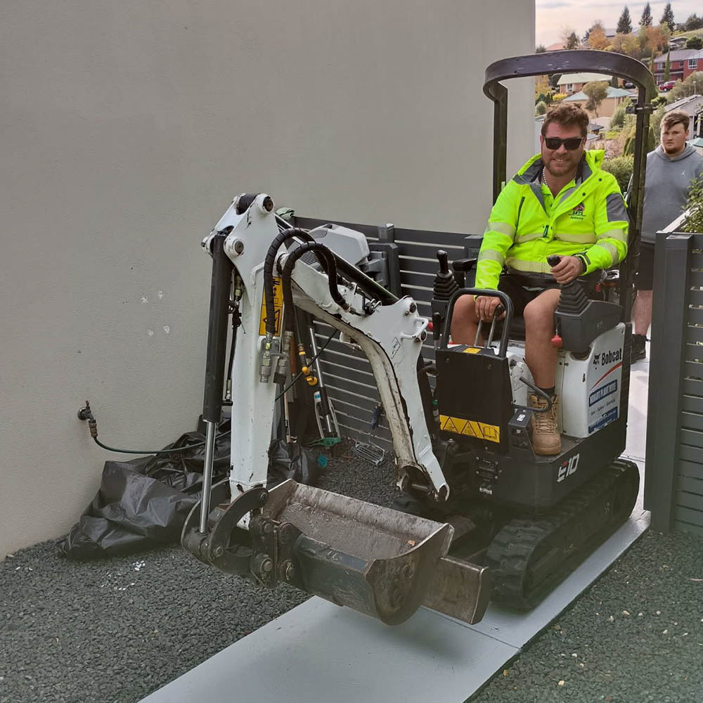 Man in hi-vis driving a mini excavator on a board beside a house