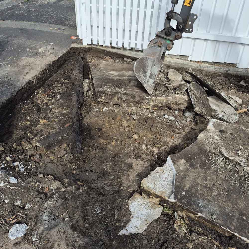 Excavator breaking up old concrete beside a white picket fence